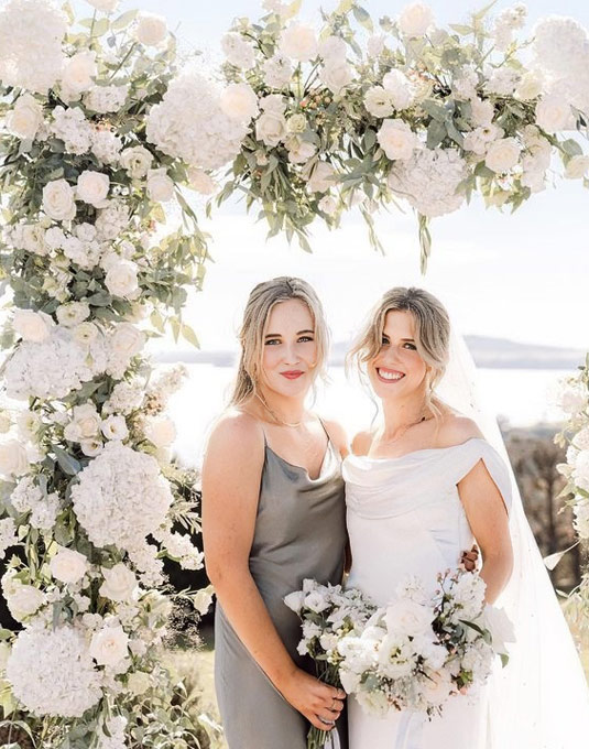 Bride and bridesmaid standing below flower arch.