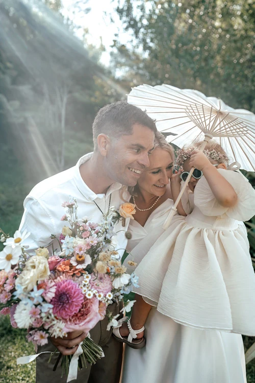 Real bride in convertible wedding dress with long skirt, groom, and flower girl under a parasol – Auckland, New Zealand.